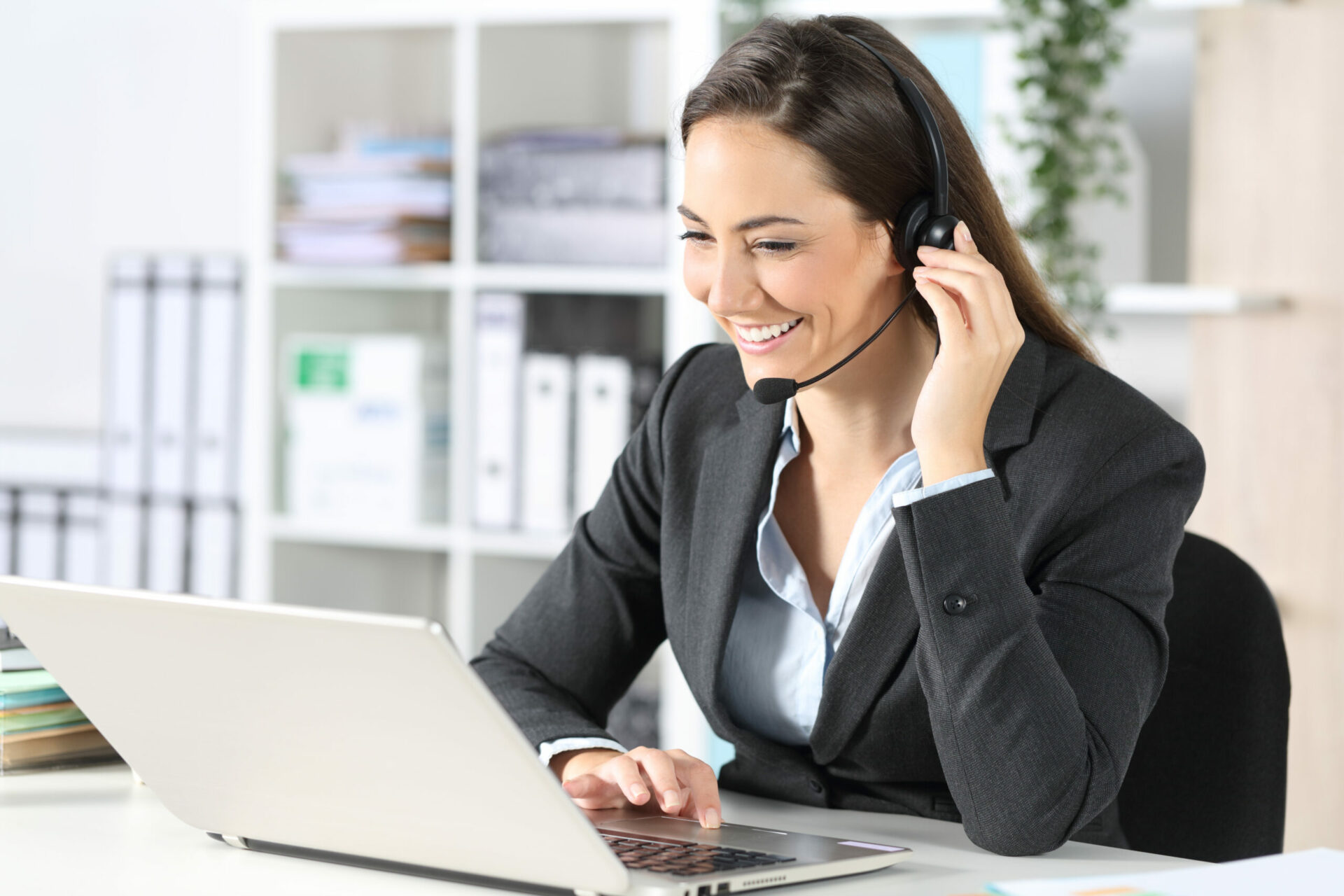 Happy telemarketer woman with laptop attending call with headset sitting on a desk at the office